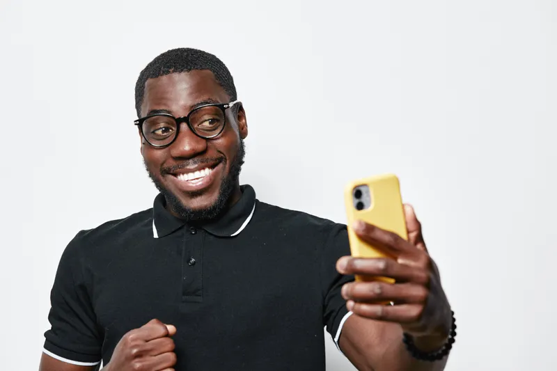 Black man with dark skin and glasses smiling while taking selfie with yellow smartphone. Casual clothing, happy expression, isolated on white background with positive mood and technology use.
