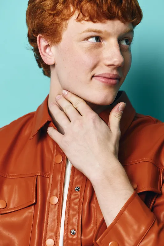 Young man with red hair wearing brown leather jacket touching neck and looking away with calm expression on blue background in studio portrait. Casual fashion and style.