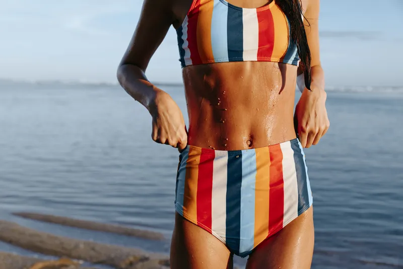 Woman bikini beach ocean striped summer sunlight, close up torso and toned skin, walking along shoreline, gentle waves, sunny outdoor mood, vibrant colors fresh sea breeze and relaxed pose