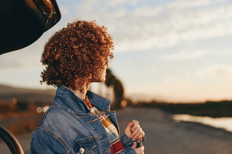 Woman lifestyle, outdoors at sunset, curly hair, denim jacket, casual jeans, she walks on a road, relaxed travel mood, holding hand with a companion, warm evening light