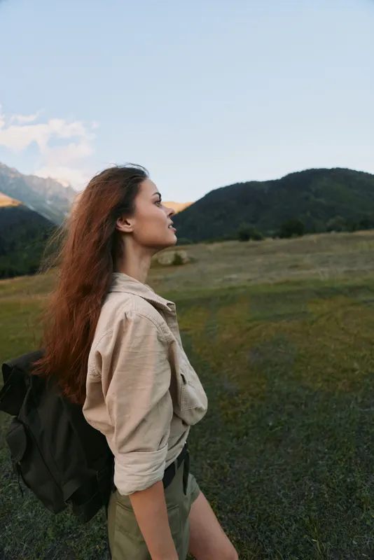 Young woman traveler with long hair and backpack enjoys mountain landscape during outdoor adventure, standing on grassy field with clear sky and hills in background.