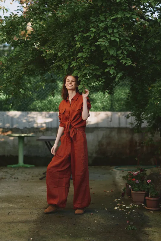Cheerful woman outdoors in a rust jumpsuit, smiling softly, enjoying a relaxed lifestyle moment amid lush trees and soft sunlight, candid portrait with natural colors and calm mood