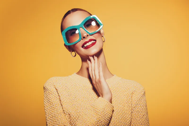Stylish woman with large turquoise sunglasses and red lipstick smiles confidently against yellow background. She wears a textured beige sweater and gold hoop earrings. Fashion and beauty concept.