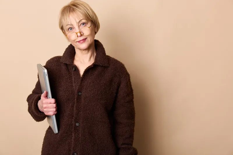 Elderly woman smiling while holding a smartphone in her hand, studio portrait on beige background, soft light, technology and communication, everyday life, calm friendly mood