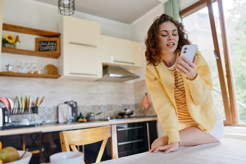 A woman in a yellow cardigan sits on a kitchen counter, looking at her phone in a bright, cozy home scene. A candid moment of modern lifestyle and daily routine.