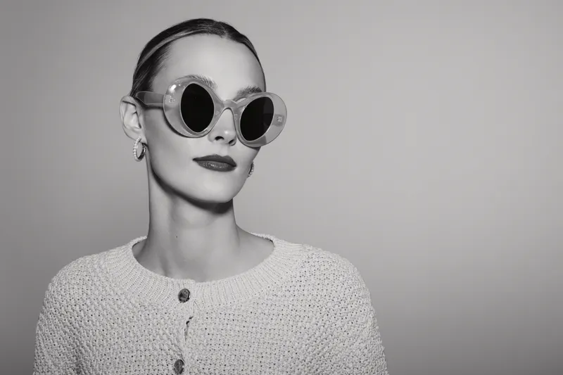 Stylish white woman with fair skin wearing round sunglasses and knitted sweater posing with confident expression on gray background.