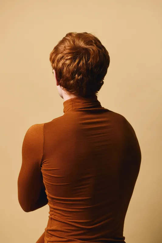 Back view of person with red hair wearing brown turtleneck sweater standing against plain beige background. Portrait, casual clothing, studio lighting and simple minimalist style.
