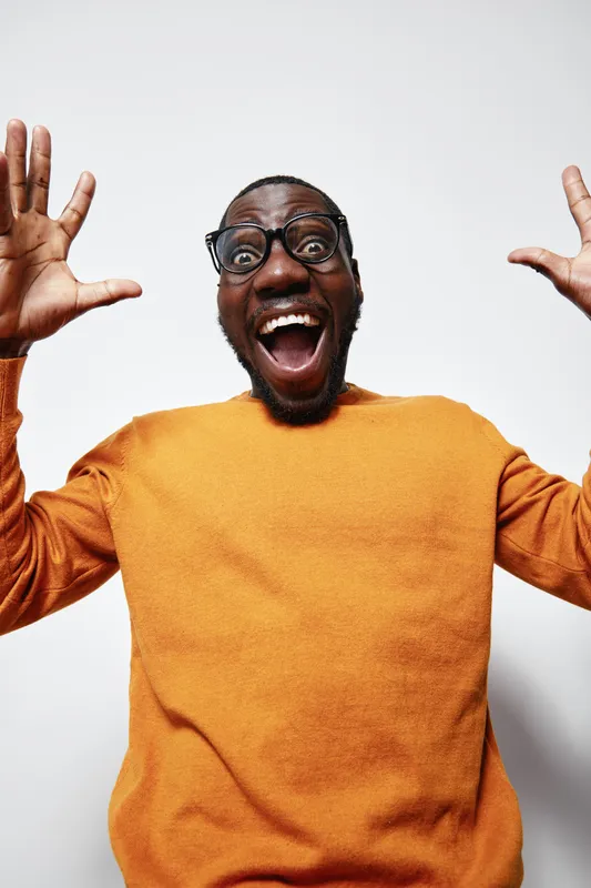 African man with dark skin and glasses wearing orange sweater expressing excitement with wide open mouth and raised hands on white background.