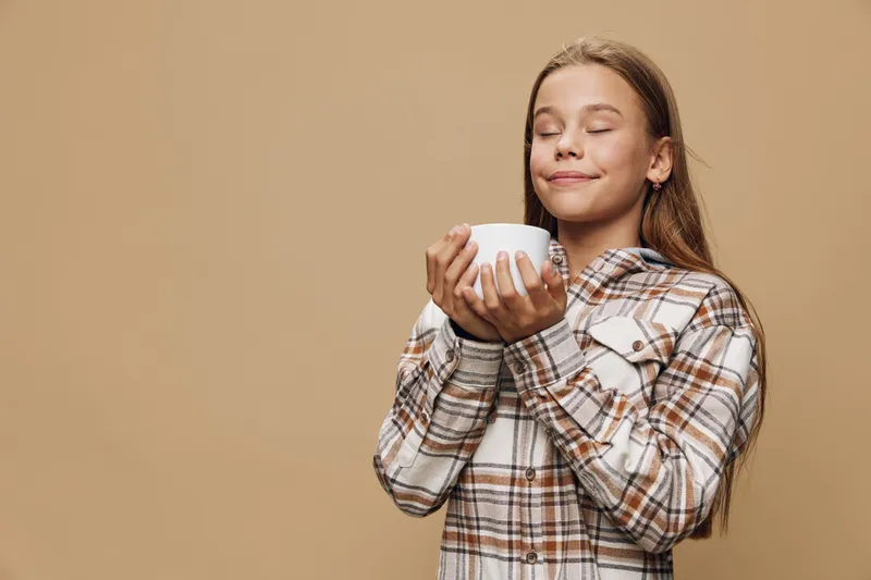 Girl with long hair in plaid shirt holding a white cup with closed eyes enjoying warm beverage, relaxed mood and cozy autumn vibe on neutral background.