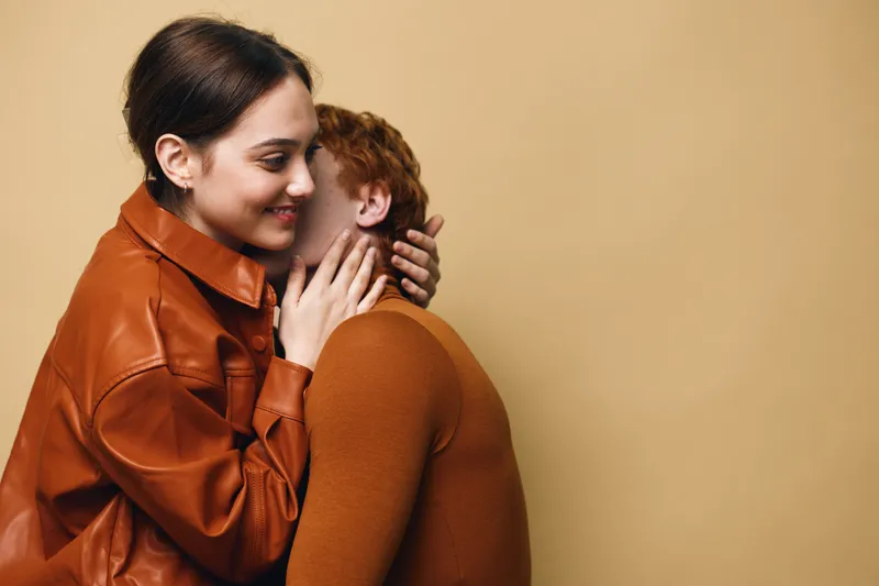 Young couple embracing indoors with warm smiles and affection. Woman in brown leather jacket hugging man wearing knitted sweater against beige background in cozy moment.