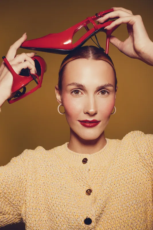 Young woman with red high heels holds shoes above her head, wearing yellow knitted sweater and gold hoop earrings. Fashion concept with confident model on brown background.