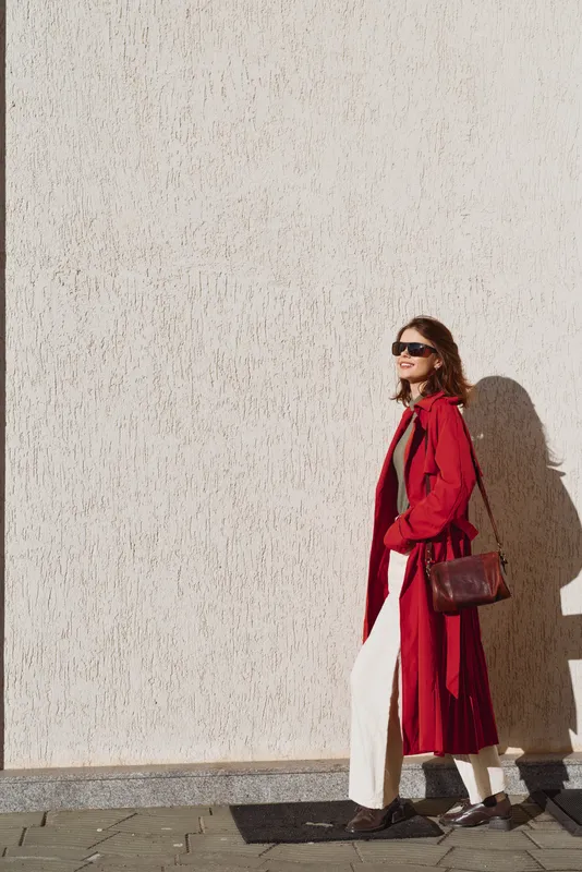 Cheerful woman outdoors wearing a red coat and white pants, sunglasses, carrying a brown bag, posing against a sunlit wall with relaxed street style and breeze