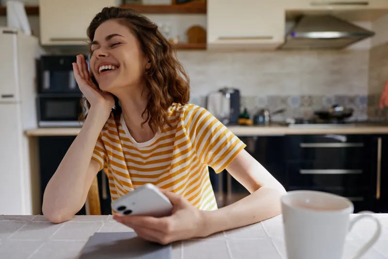 A smiling woman sits at a bright kitchen table, holding a phone, enjoying a candid home moment. Casual lifestyle scene highlighting everyday joy, connection, and comfort in a warm domestic setting.