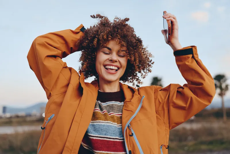 Woman with curly hair, smiling outdoors, wearing orange jacket, holding a phone in hand, playful travel lifestyle, sunny sky background, wind in hair, candid street moment