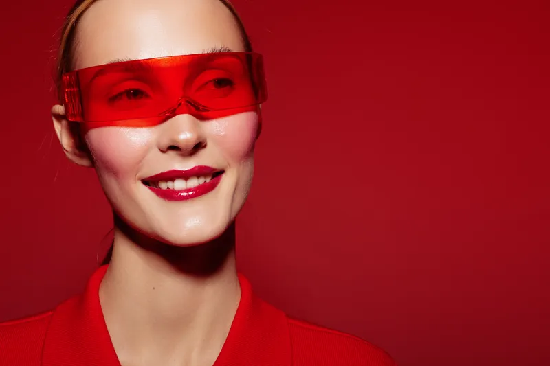 Smiling woman in red outfit and red transparent glasses poses on red background. Concept of fashion, style, modern accessories, and confident female model with bright makeup.