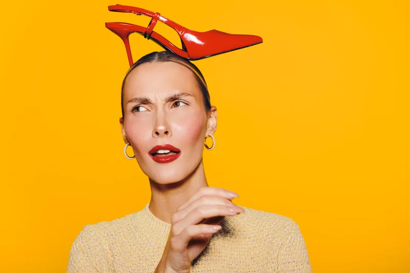 Confused woman with red lipstick and gold earrings holds red high heel shoe on head against bright yellow background. Concept for fashion, style, and quirky emotions.