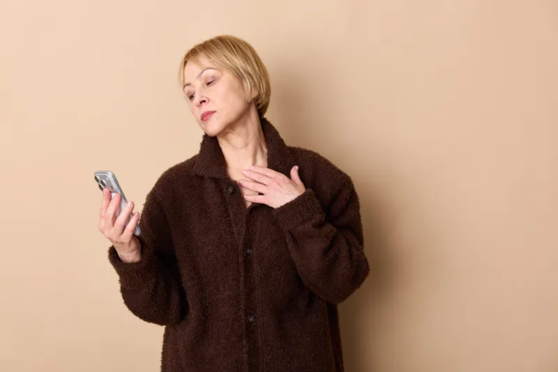 Woman with smartphone indoors on beige background wearing dark fuzzy jacket, worried expression, touching neck, suggesting cold weather, soft studio light