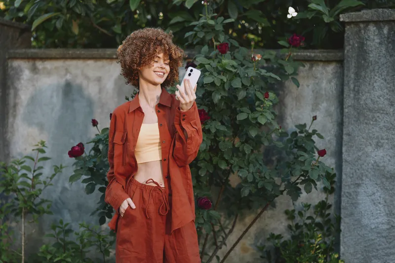 A stylish woman in an orange rust outfit takes a casual selfie in a sunlit garden, posing beside a stone wall. Fresh lifestyle portrait with cheerful mood and natural backdrop