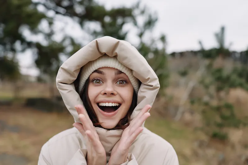 Woman outdoors in a warm winter coat and beanie, excited smile, framing face with hands, natural garden setting, cozy apparel and candid joyful energy.