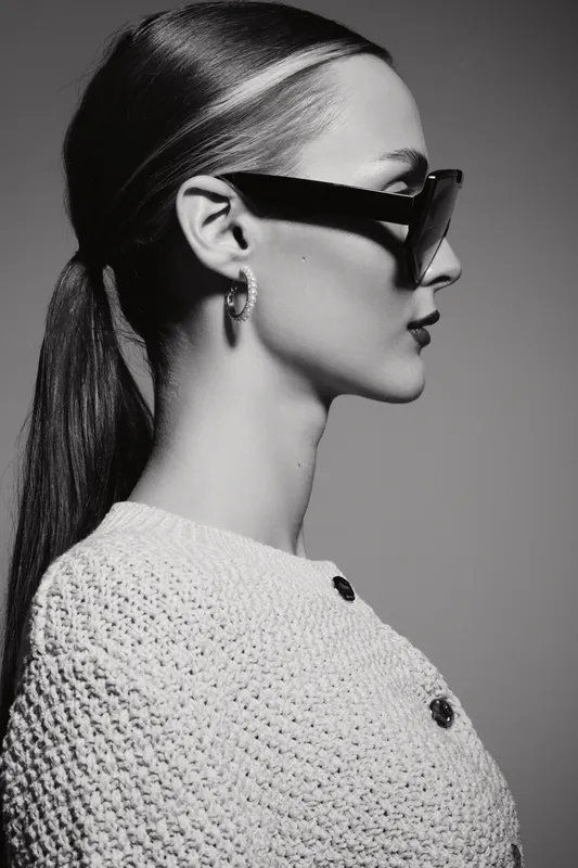 Profile of a young Caucasian woman with light skin wearing sunglasses and knitted sweater in a studio portrait.