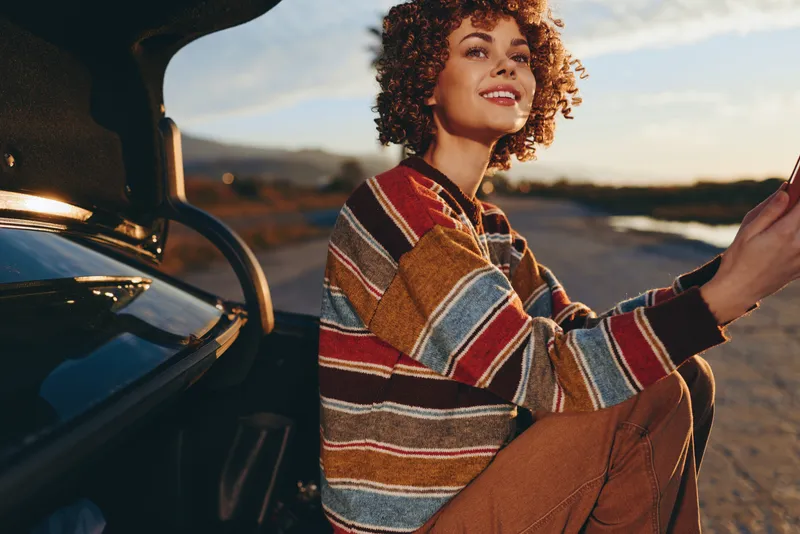 Woman lifestyle smiling at sunset while driving a car on an open road, hands on steering wheel, wearing a striped sweater, warm golden light, outdoor travel moment