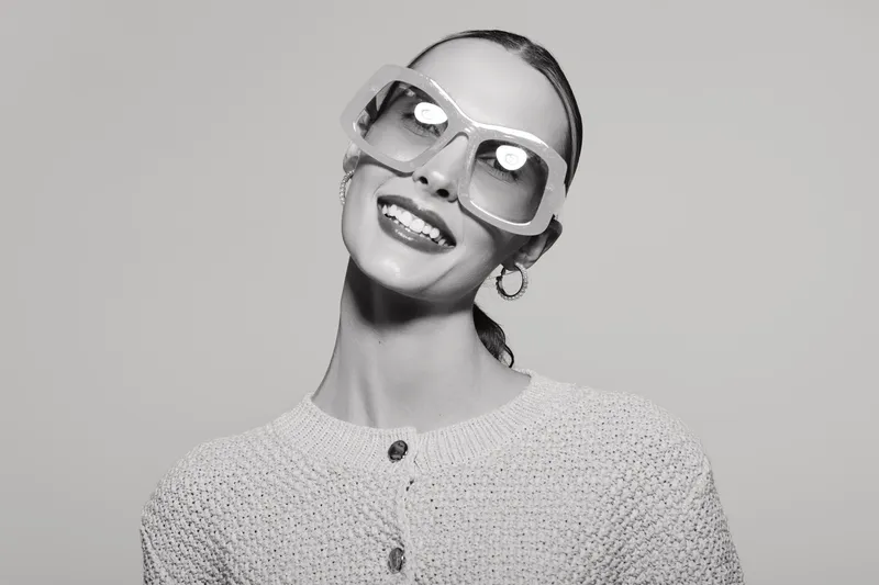 Smiling white woman with fair skin and slicked hair wears oversized sunglasses and knitted sweater, posing against grey background.