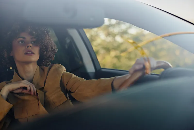 A woman driver in retro clothing sits behind the wheel, gazing ahead with a calm expression. The interior is vintage and softly lit, highlighting lifestyle, travel, and daytime city moments.