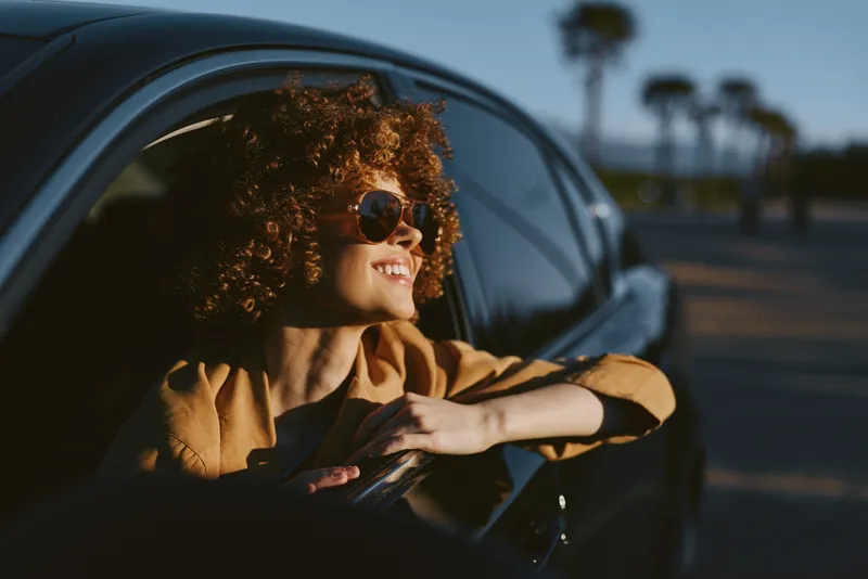Vibrant woman lifestyle outdoors, smiling from a car window, sunglasses on, sunlit breeze, retro film color palette, candid and relaxed moment