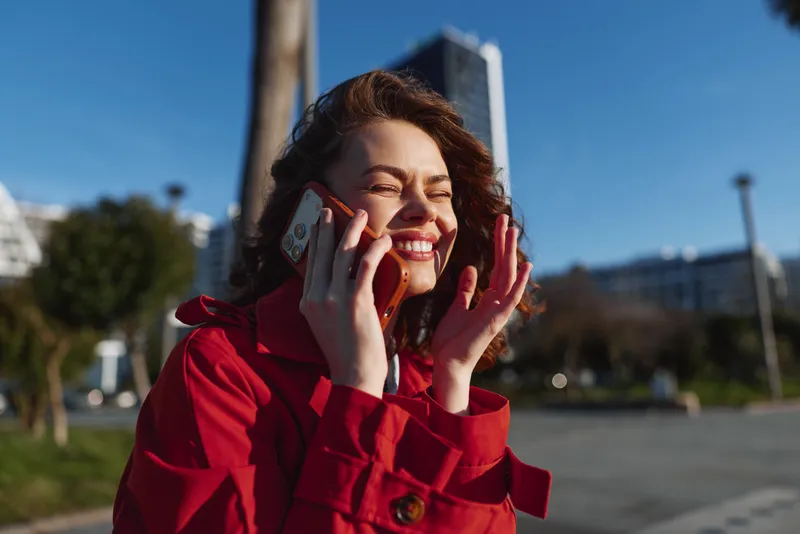 Smiling young woman in a red coat talks on the phone in an outdoor urban portrait, a candid phone conversation capturing brunette female confidence in bright afternoon light.