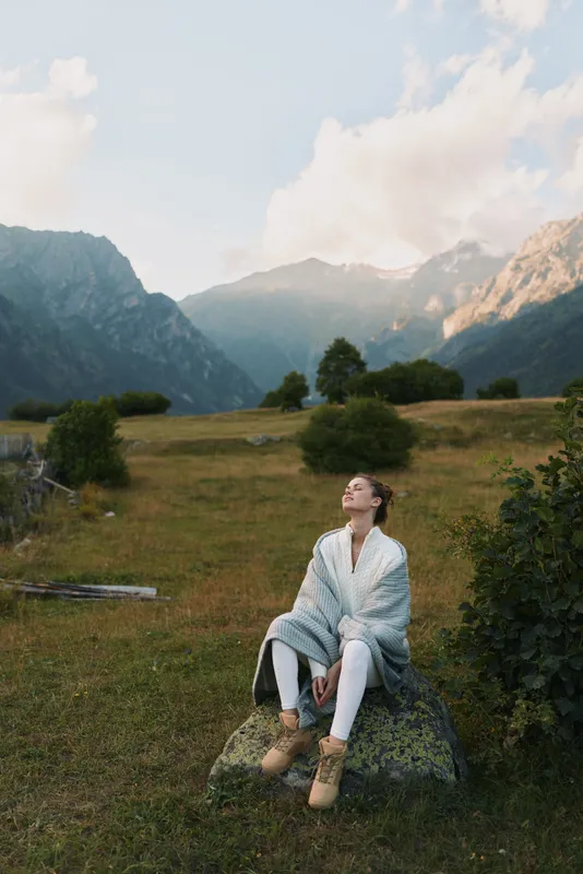 Young woman sits on a rock in a grassy meadow with distant mountains, savoring a peaceful outdoor moment, scene filled with soft light, warm tones and quiet contemplation