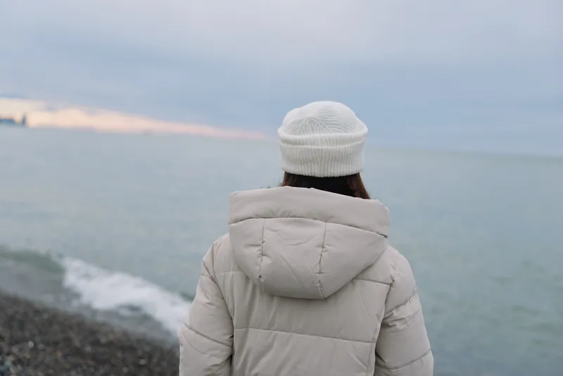 Winter scene with a person in a light jacket and beanie facing the sea at the coast, calm horizon, soft light, reflective mood, outdoor solitude