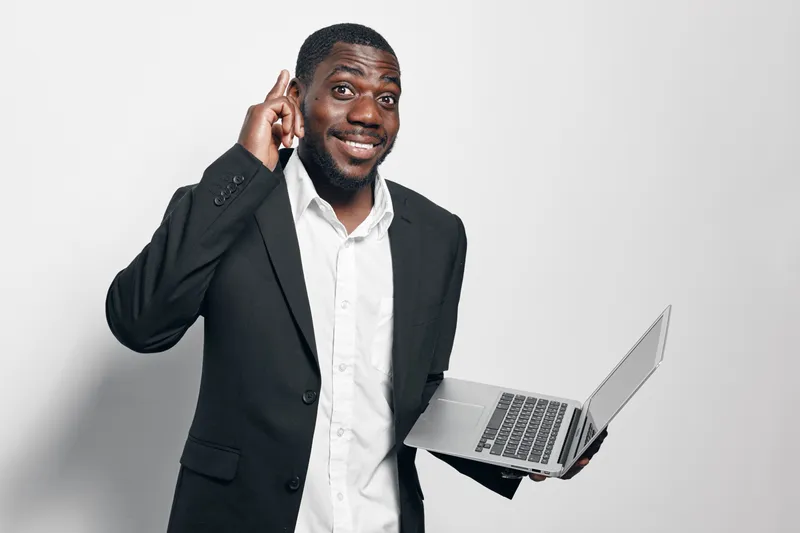 African American man in black suit holding laptop and pointing finger up with smile on white background. Business professional with dark skin and confident expression in studio.