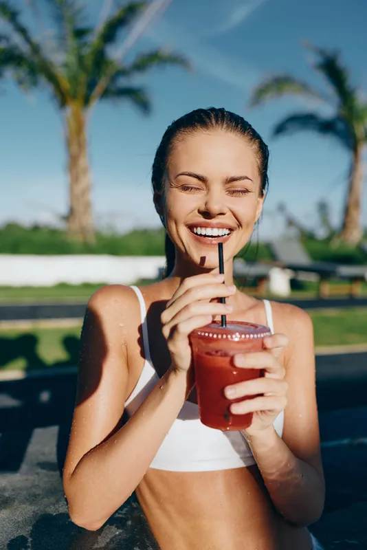 A sunlit poolside scene shows a young woman in a white sports bra smiling as she sips a red juice through a straw, with palm trees in the background.