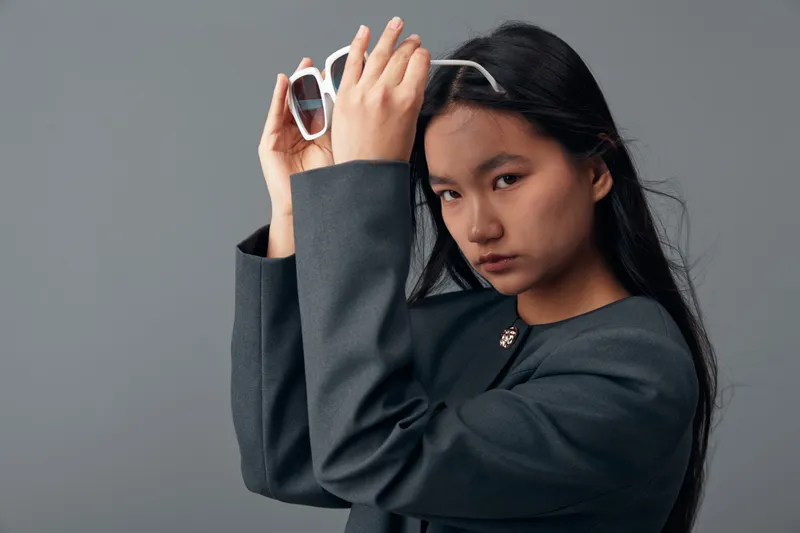 Young woman with long black hair holding white sunglasses wearing dark oversized blazer looking confidently at camera with gray background. Fashion, portrait and style concept.