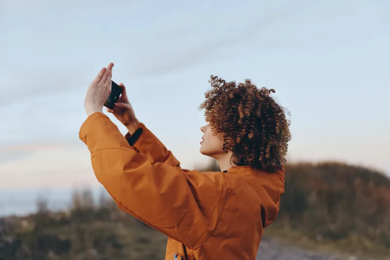 Woman lifestyle outdoors, curly hair, orange jacket, raises hand for playful pose, sunset portrait, calm expression, looking to the horizon, natural landscape background