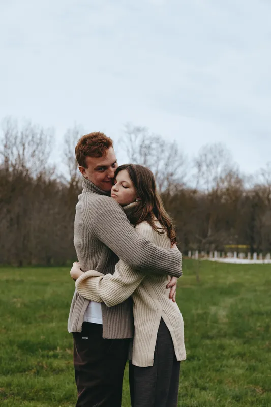 A young couple embraces tenderly outdoors on a cloudy day as they wear cozy knitted sweaters in a quiet natural setting with bare trees and green grass around them.