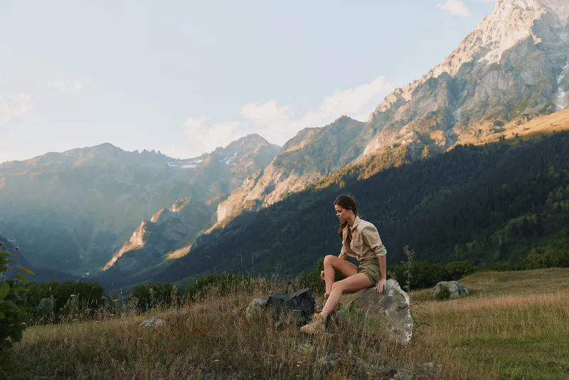 Young woman sitting on a rock in a tranquil meadow with towering mountains behind, enjoying a peaceful moment outdoors and soaking in fresh air.