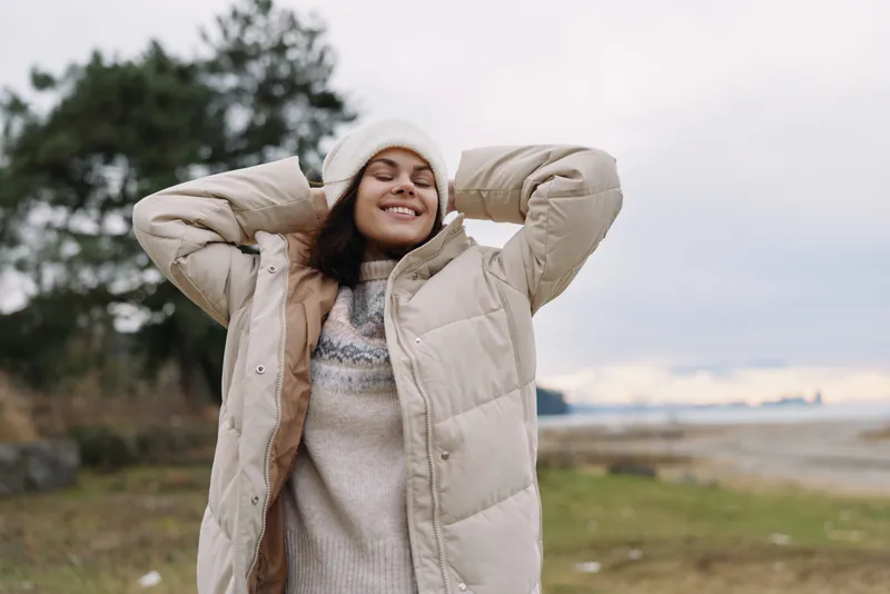 Woman outdoors wearing a warm beige jacket, smiling with hands behind her head, enjoying chilly weather near a coastline, relaxed pose, cozy attire, natural light and wind
