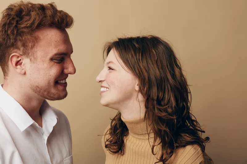 Smiling young couple looking at each other with affection, standing close on beige background. Concept of love, relationship, happiness, and connection between people.