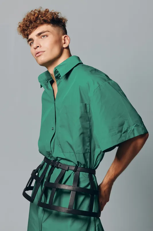 Young man with short curly hair in green oversized shirt and wide belt posing against gray background in studio. Male fashion model shows modern streetwear style and casual outfit profile.
