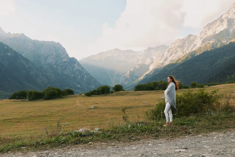 Young woman in light sweater stands alone in a wide mountain valley enjoying peaceful nature and bright morning light