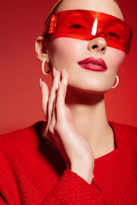 Stylish woman in red sweater and futuristic red sunglasses poses with hand near face on red background. Concept of fashion, beauty, modern style, and confident attitude in studio lighting.