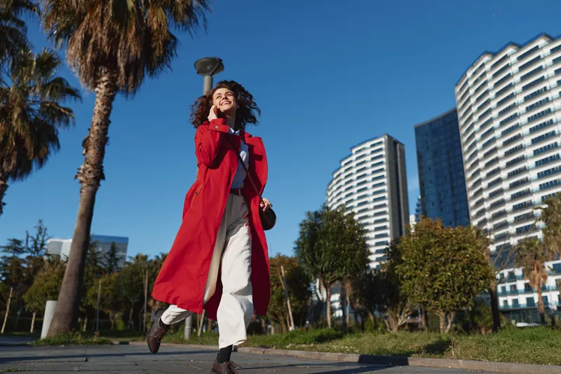 A woman in a long red coat walks along a sunny urban street, her confident stride framed by modern buildings and palm trees, capturing bold street style in motion.