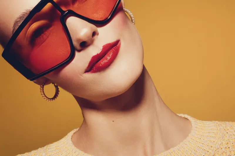 Close-up of stylish woman wearing oversized red sunglasses and gold hoop earrings on yellow background. Fashion portrait with bold makeup and trendy accessories.