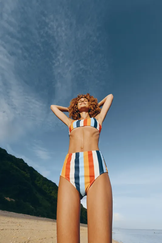 Woman in bikini on beach posing with hands behind head under summer sunshine, ocean horizon and blue sky behind, mountains at left, outdoor vacation mood, natural light, curly hair