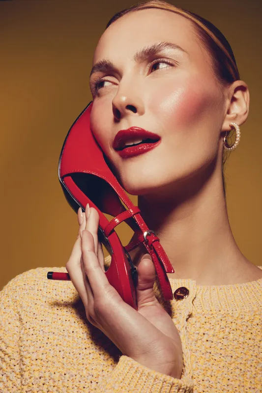 Close-up of a fashionable woman holding a red high heel shoe near her face. She wears bold makeup and a yellow sweater, posing against a warm brown background. Concept of style.