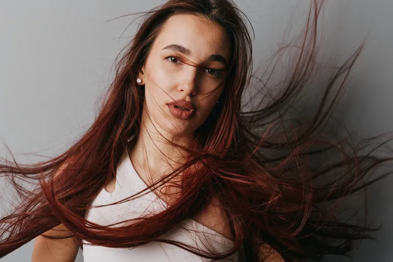 Woman with long brown hair blowing in wind wearing white top with natural makeup and pearl earrings posing against gray background with soft lighting and expressive face.
