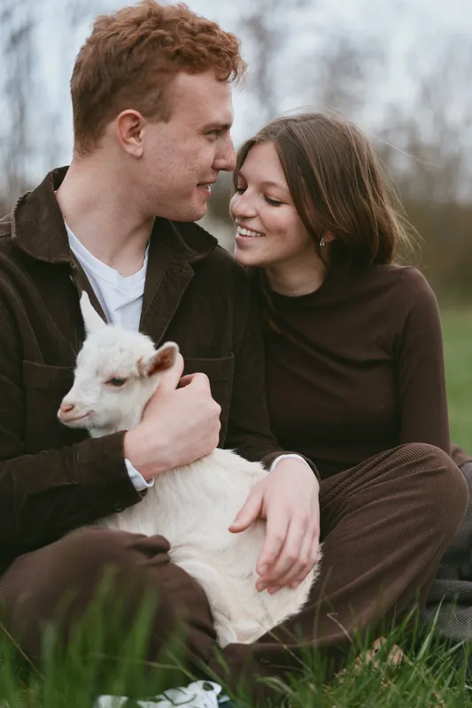 Couple shares a gentle moment outdoors with a young goat kid, smiling softly in a grassy meadow, showcasing rustic lifestyle, affection, companionship, and natural light in a peaceful rural setting.