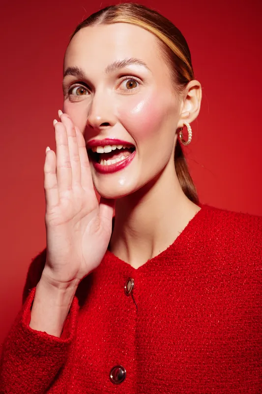 Young woman in red sweater whispers with hand near mouth against red background. Concept of secret, communication, and expression with bright studio lighting and close-up portrait.