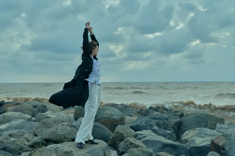 Woman stands on jagged rocks by the sea, arms raised under a windy, overcast sky, wearing a long coat and light trousers, capturing a moment of freedom and resilience against coastal elements.